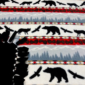 May include: White fleece fabric with a repeating pattern of black bears, foxes, and birds flying over a mountain range with pine trees. The fabric has a red, white, and brown color scheme.