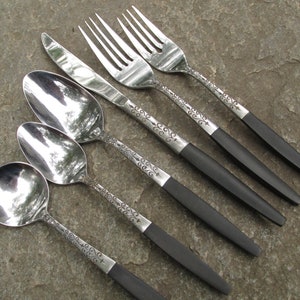 May include: A set of five silver-toned flatware pieces with black handles. The set includes a knife, two forks, and two spoons. The handles have a decorative floral pattern.