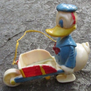 May include: A vintage Donald Duck figurine pushing a small wheelbarrow. The figure is wearing a blue sailor suit, a red bow tie, and a blue hat. The wheelbarrow is red, blue, and yellow. The background is a gray, textured surface.