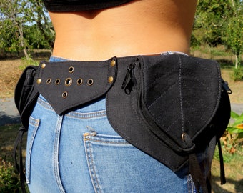 May include: Black fanny pack with multiple compartments and a leaf-shaped design. The pack features brass-colored grommets and zippers. It is worn around the waist over blue jeans.