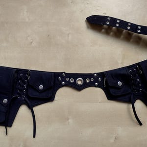 Op de afbeelding: Zwarte tailleband met meerdere zakken en zilverkleurige metalen accenten. De riem heeft een gespsluiting, oogjes en decoratieve klinknagels. Het ontwerp omvat vetersluiting en een centrale metalen ring.