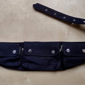 Könnte beinhalten: Eine schwarze Bauchtasche mit drei Reißverschlusstaschen und silberfarbenen Beschlägen. Der verstellbare Gürtel hat eine Metallschnalle. Der Gürtel ist ca. 102 cm lang.