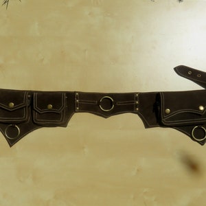 Può includere: Una cintura in pelle marrone con più tasche e anelli metallici. La cintura ha un dettaglio di cucitura decorativo.