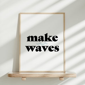May include: A framed print with the text "make waves" in black bold font with a blue wave graphic above the word "waves". The print is on a light wood shelf against a white wall.