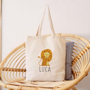 May include: A beige canvas tote bag with a lion illustration and the name "Luca" printed on the front. The bag is sitting on a wicker chair.
