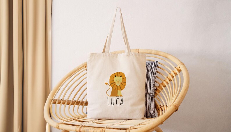 May include: A beige canvas tote bag with a lion illustration and the name "Luca" printed on the front. The bag is sitting on a wicker chair.
