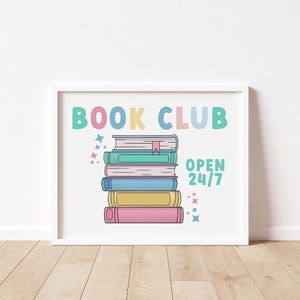 K&ouml;nnte beinhalten: Wei&szlig;er gerahmter Druck mit dem Text "BOOK CLUB" in bunten Buchstaben. Unter dem Text ist ein Stapel bunter B&uuml;cher mit einem Lesezeichen abgebildet. Der Text "OPEN 24/7" befindet sich unter den B&uuml;chern.