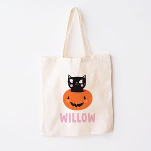 May include: A white canvas tote bag with a black cat inside a jack-o'-lantern. The bag has the name "WILLOW" printed on the front in pink.