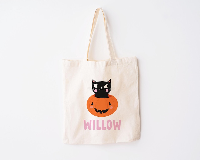May include: A white canvas tote bag with a black cat inside a jack-o'-lantern. The bag has the name "WILLOW" printed on the front in pink.