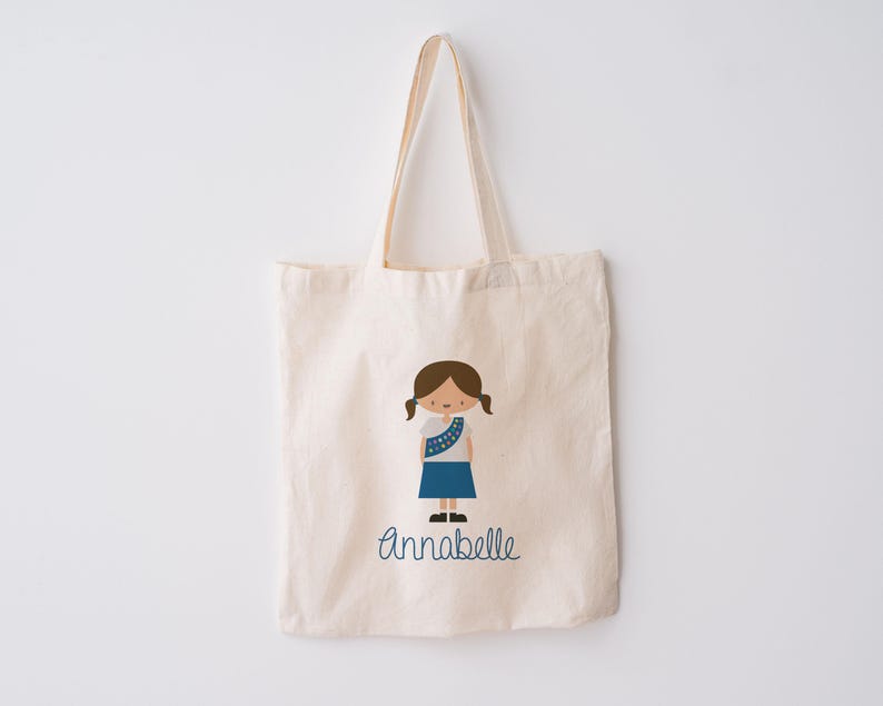 May include: A white canvas tote bag with a blue and brown illustration of a girl scout. The bag has the name "Annabelle" printed in blue cursive.