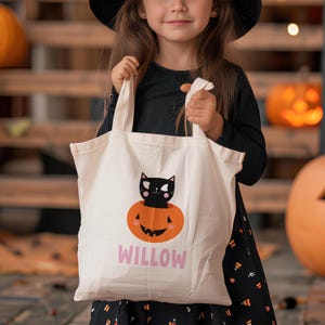 May include: A white canvas tote bag with a black cat inside an orange pumpkin. The pumpkin has a smiling face. The bag has the name "WILLOW" printed in pink letters.