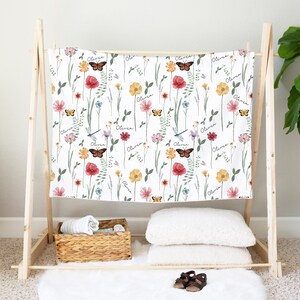 May include: A white baby blanket with a floral pattern featuring butterflies, dragonflies, and the name "Olivia" repeated throughout. The blanket is displayed on a wooden stand.