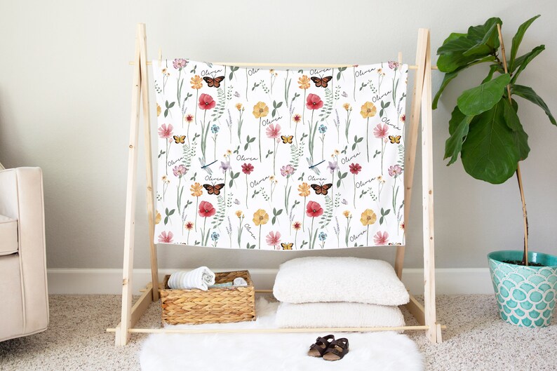 May include: A white baby blanket with a floral pattern featuring butterflies, dragonflies, and the name "Olivia" repeated throughout. The blanket is displayed on a wooden stand.