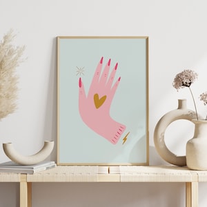 Può includere: Una stampa incorniciata di una mano rosa con un cuore dorato sul palmo, smalto per unghie rosa e un fulmine dorato sul polso. Lo sfondo è azzurro con una stella dorata.