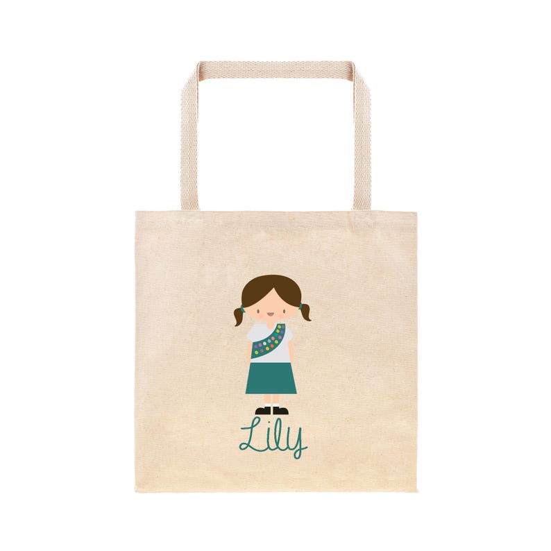 May include: A beige tote bag featuring a cartoon illustration of a girl scout wearing a green sash and a white shirt. The girl has brown hair and is smiling. The text "Lily" is written in green below the illustration.