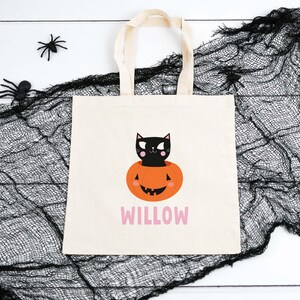 May include: A white canvas tote bag with a black cat sitting inside a jack-o'-lantern. The bag has the name "WILLOW" printed in pink letters.