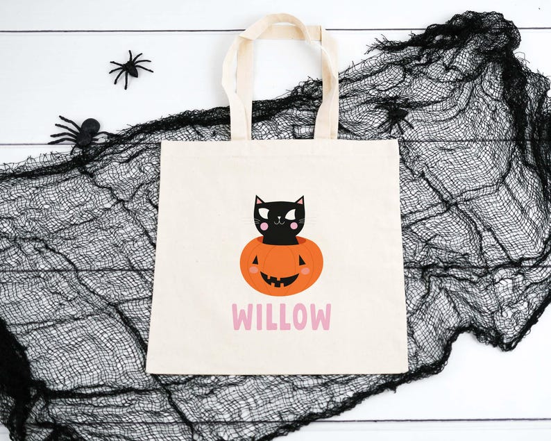 May include: A white canvas tote bag with a black cat sitting inside a jack-o'-lantern. The bag has the name "WILLOW" printed in pink letters.
