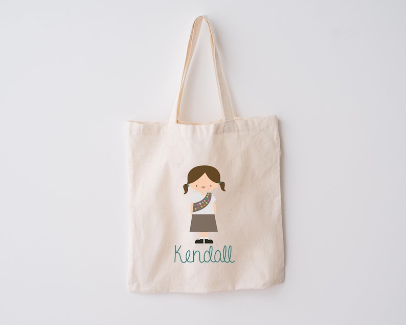 May include: A white canvas tote bag featuring a brown-haired girl in a scout uniform with the name "Kendall" printed in teal.