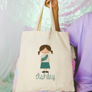 May include: A white canvas tote bag featuring a green illustration of a girl scout and the name "Ashley" written in green.