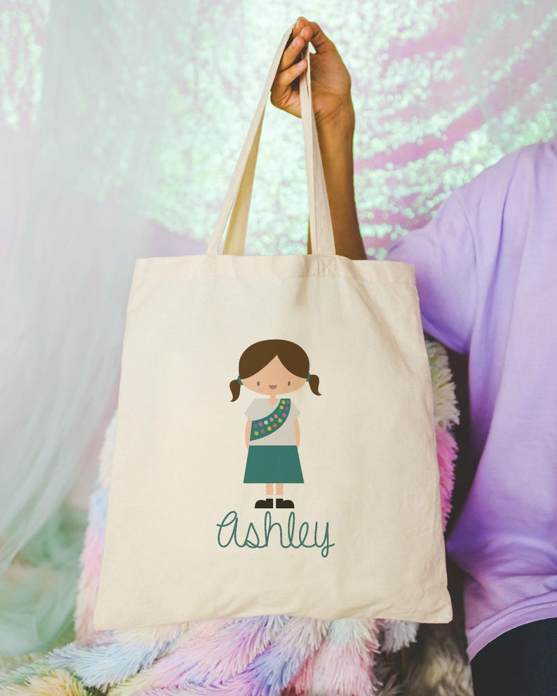 May include: A white canvas tote bag featuring a green illustration of a girl scout and the name "Ashley" written in green.