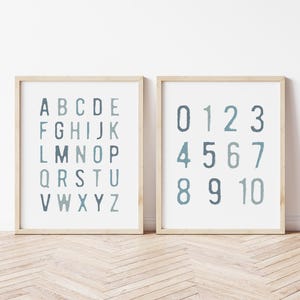 May include: Two framed prints with a light wood finish. One print displays the alphabet in uppercase letters, and the other displays the numbers 0 to 10. The letters and numbers are in a blue-green watercolour style.
