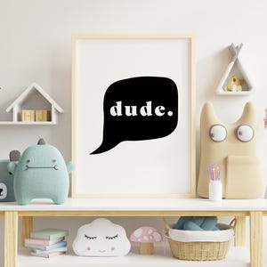 May include: Black and white print with the word "dude" in a speech bubble. The print is framed and hanging on a white wall above a wooden shelf with toys and decor.