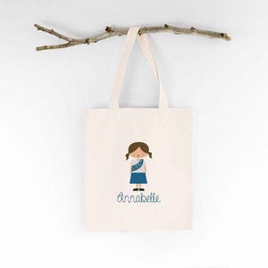 May include: A white canvas tote bag with a blue and white illustration of a girl wearing a blue uniform and a blue sash. The text "Annabelle" is printed below the illustration.