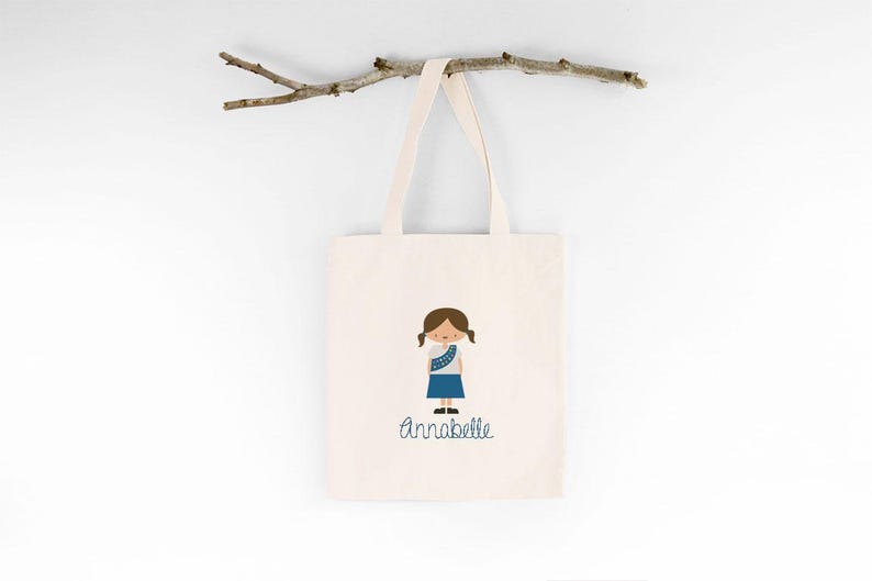May include: A white canvas tote bag with a blue and white illustration of a girl wearing a blue uniform and a blue sash. The text "Annabelle" is printed below the illustration.
