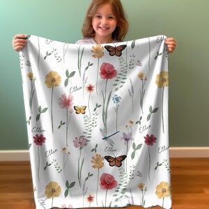 May include: A white baby blanket with a floral pattern featuring butterflies, dragonflies, and the name "Ellie" repeated throughout. The blanket is soft and cuddly, perfect for a baby or toddler.
