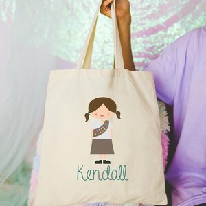 May include: A white canvas tote bag featuring a cartoon illustration of a girl guide wearing a brown uniform and a sash with colourful badges. The text "Kendall" is printed in green below the illustration.