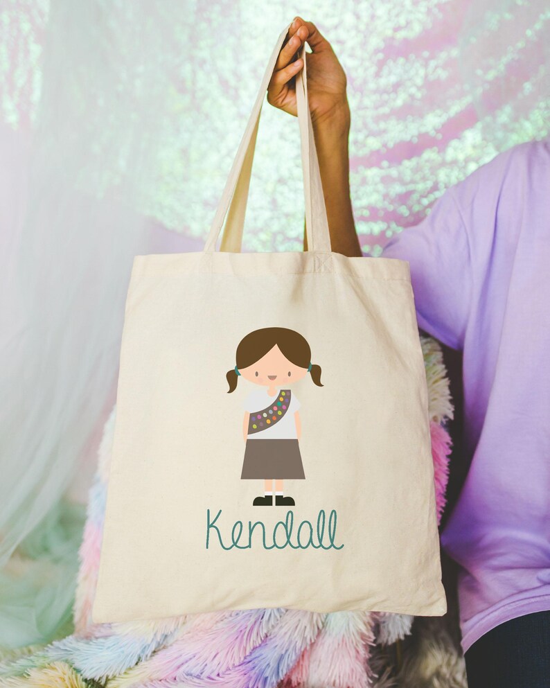 May include: A white canvas tote bag featuring a cartoon illustration of a girl guide wearing a brown uniform and a sash with colourful badges. The text "Kendall" is printed in green below the illustration.