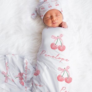 May include: A white swaddle blanket with a pink cherry print and the name "Penelope" written in pink script. A baby is wearing the swaddle and a matching pink cherry print hat.
