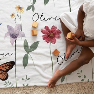 May include: A white baby blanket with a floral print featuring butterflies, dragonflies, and the name "Olivia" written in cursive. The blanket is on a carpeted floor with a child playing with wooden blocks.