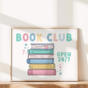 K&ouml;nnte beinhalten: Ein wei&szlig;er gerahmter Poster mit dem Text "BOOK CLUB" in bunten Buchstaben. In der Mitte des Posters ist ein Stapel von f&uuml;nf B&uuml;chern abgebildet. Der Text "OPEN 24/7" befindet sich unter den B&uuml;chern.