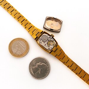 K&ouml;nnte beinhalten: Eine goldfarbene Armbanduhr mit einem rechteckigen Zifferblatt und einem goldfarbenen Metallband. Die Uhr liegt auf einer wei&szlig;en Oberfl&auml;che mit einer Euro-M&uuml;nze und einem US-amerikanischen Viertel neben ihr. Das Zifferblatt der Uhr ist ge&ouml;ffnet und zeigt das Innenleben der Uhr.