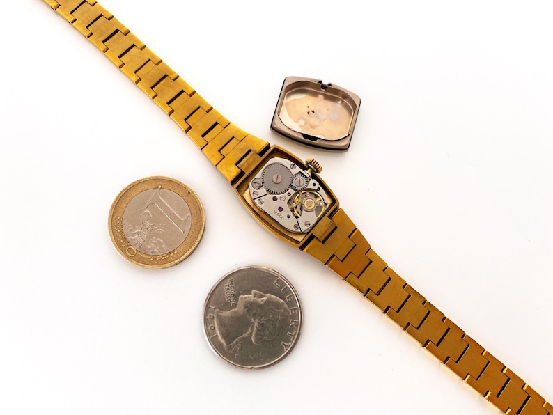 K&ouml;nnte beinhalten: Eine goldfarbene Armbanduhr mit einem rechteckigen Zifferblatt und einem goldfarbenen Metallband. Die Uhr liegt auf einer wei&szlig;en Oberfl&auml;che mit einer Euro-M&uuml;nze und einem US-amerikanischen Viertel neben ihr. Das Zifferblatt der Uhr ist ge&ouml;ffnet und zeigt das Innenleben der Uhr.
