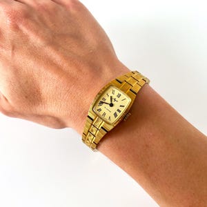 K&ouml;nnte beinhalten: Eine goldfarbene Armbanduhr mit quadratischem Zifferblatt und r&ouml;mischen Ziffern f&uuml;r die Stunden. Die Uhr hat ein goldfarbenes Metallband.