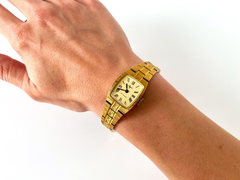 K&ouml;nnte beinhalten: Eine goldfarbene Armbanduhr mit quadratischem Zifferblatt und r&ouml;mischen Ziffern f&uuml;r die Stunden. Die Uhr hat ein goldfarbenes Metallband.
