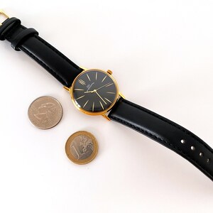 May include: A gold-tone wristwatch with a black face and black leather band. The watch is sitting on a white surface with two coins.