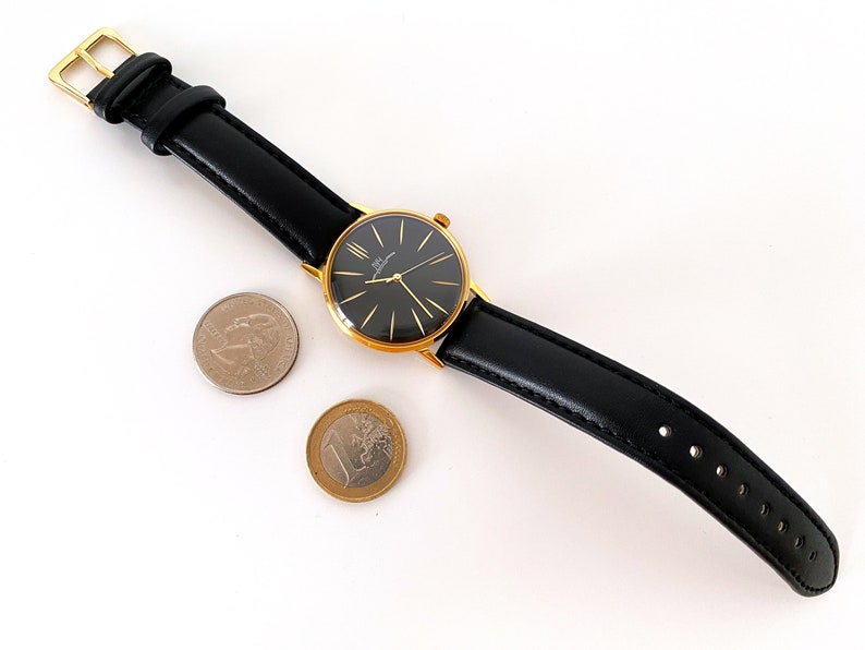 May include: A gold-tone wristwatch with a black face and black leather band. The watch is sitting on a white surface with two coins.