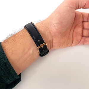 May include: A black leather watch strap with a gold buckle. The strap is worn on a person's wrist.