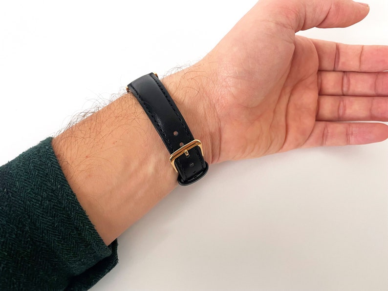 May include: A black leather watch strap with a gold buckle. The strap is worn on a person's wrist.