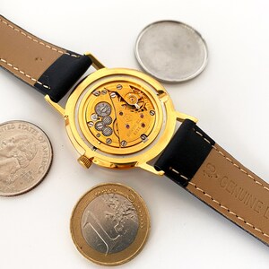 May include: A gold-tone wristwatch with a black leather band. The watch is open, revealing the inner workings of the watch mechanism. The watch is surrounded by three coins, including a US quarter and a Euro coin.