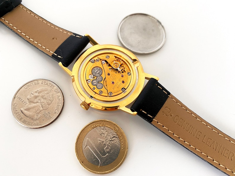 May include: A gold-tone wristwatch with a black leather band. The watch is open, revealing the inner workings of the watch mechanism. The watch is surrounded by three coins, including a US quarter and a Euro coin.