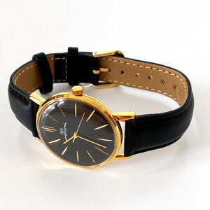 May include: A gold-tone wristwatch with a black face and black leather band. The watch face has a white dial with black Roman numeral hour markers and a white second hand.