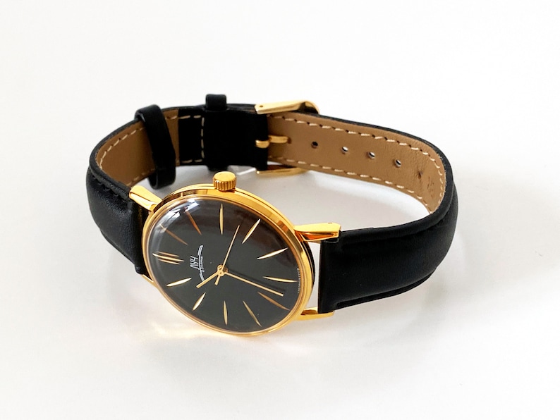 May include: A gold-tone wristwatch with a black face and black leather band. The watch face has a white dial with black Roman numeral hour markers and a white second hand.