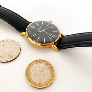 May include: A gold-tone wristwatch with a black face and black leather band. The watch is next to a US quarter and a Euro coin.