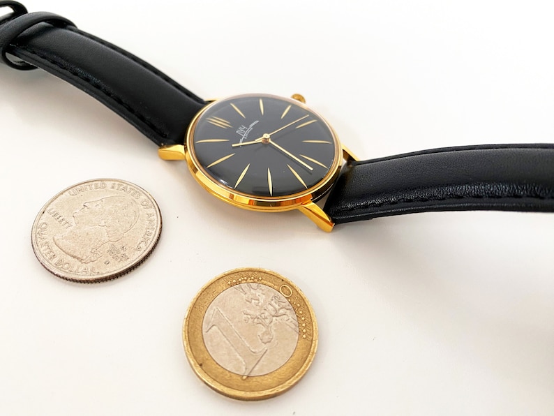 May include: A gold-tone wristwatch with a black face and black leather band. The watch is next to a US quarter and a Euro coin.