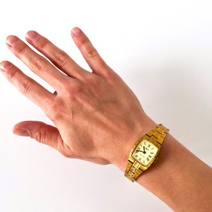 K&ouml;nnte beinhalten: Eine goldfarbene Armbanduhr mit quadratischem Zifferblatt und einem Metallband. Die Uhr hat ein wei&szlig;es Zifferblatt mit schwarzen r&ouml;mischen Ziffern als Stundenmarkierungen und schwarzen Zeigern.