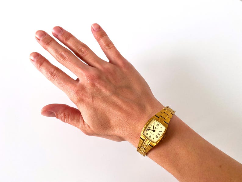K&ouml;nnte beinhalten: Eine goldfarbene Armbanduhr mit quadratischem Zifferblatt und einem Metallband. Die Uhr hat ein wei&szlig;es Zifferblatt mit schwarzen r&ouml;mischen Ziffern als Stundenmarkierungen und schwarzen Zeigern.